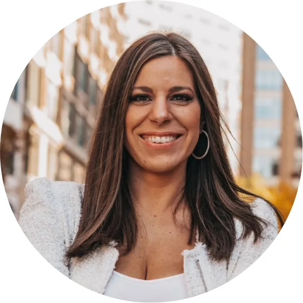 A woman with straight brown hair, wearing hoop earrings and a light-colored jacket, smiles confidently while standing outdoors in an urban setting with tall buildings in the background.
