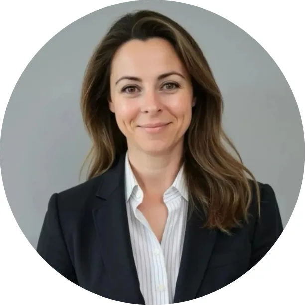 A woman with long brown hair wearing a dark blazer over a white collared shirt, smiling, posed against a plain gray background.