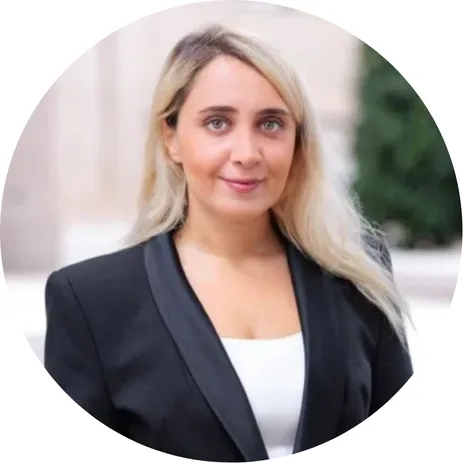A woman with long blonde hair wearing a black blazer over a white top is standing outdoors. She is looking at the camera and smiling softly. The background is blurred with greenery and a light-colored building.