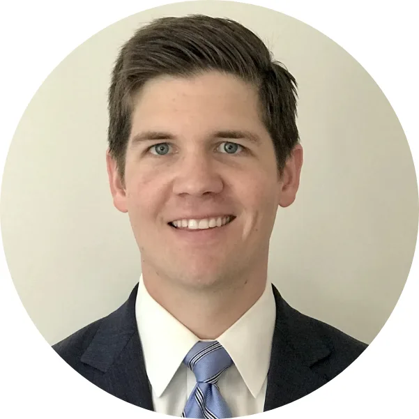 A man with short brown hair, wearing a dark suit, white shirt, and striped blue tie, smiles at the camera against a light plain background.