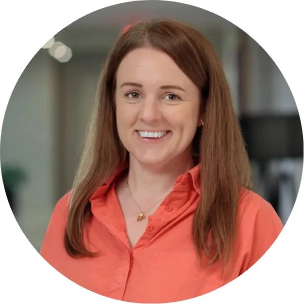 A woman with long brown hair wearing a coral button-up shirt smiles at the camera. The background is softly blurred, suggesting an indoor setting.