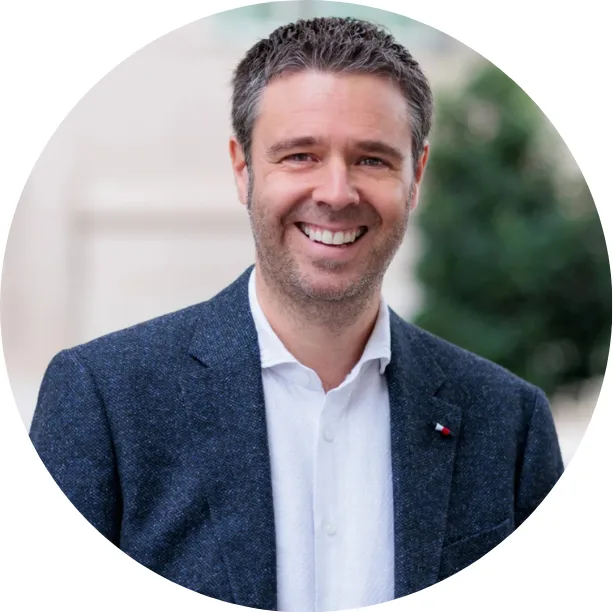 A man with short brown hair and a beard is smiling, wearing a white shirt and a dark blazer, standing outdoors with blurred greenery in the background.