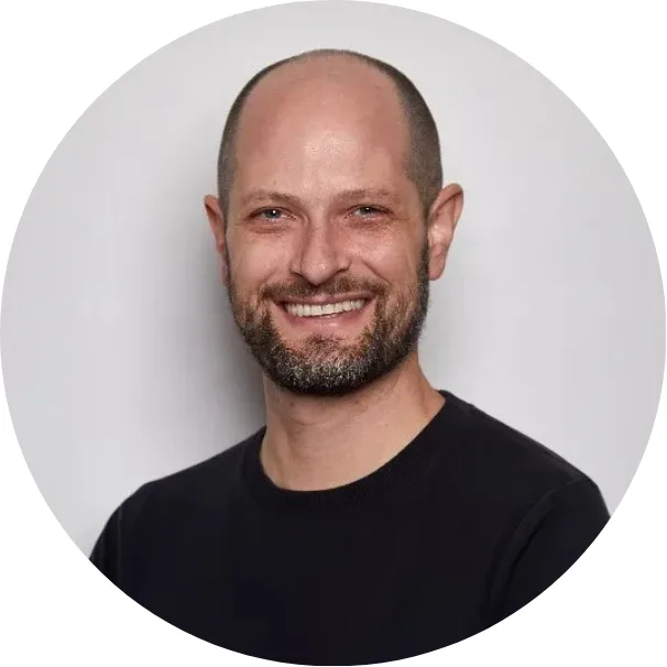 A smiling man with a bald head and short beard, wearing a black crew-neck shirt, poses against a plain light gray background.