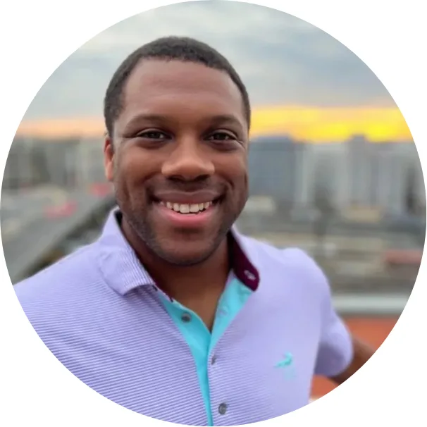 A man smiles at the camera while standing outdoors with a cityscape and sunset in the background. He is wearing a light purple collared shirt with aqua accents.