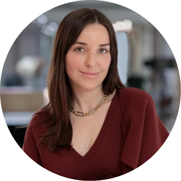 A woman with long brown hair wearing a maroon top and a beaded necklace smiles softly. The background is blurred, showing an indoor setting with soft lighting.