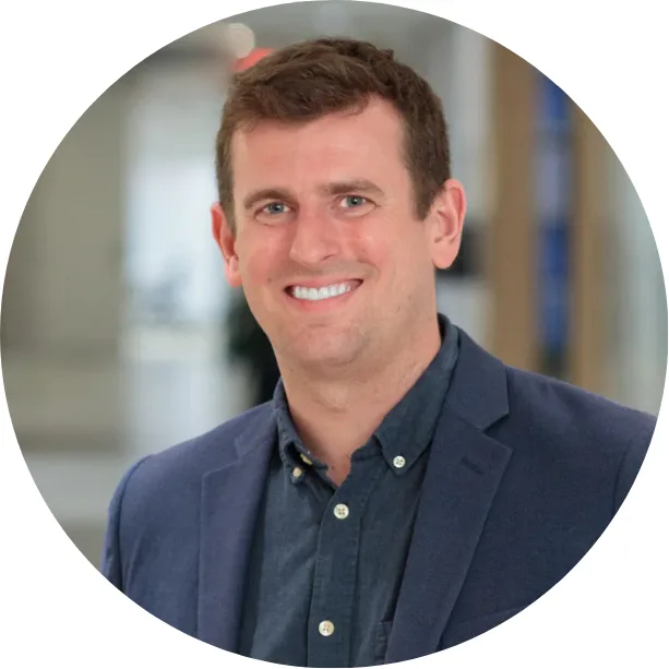 A smiling man with short brown hair, wearing a navy blue blazer over a dark shirt, stands in a bright indoor setting. The background is softly blurred, and the image is framed in a circular crop.