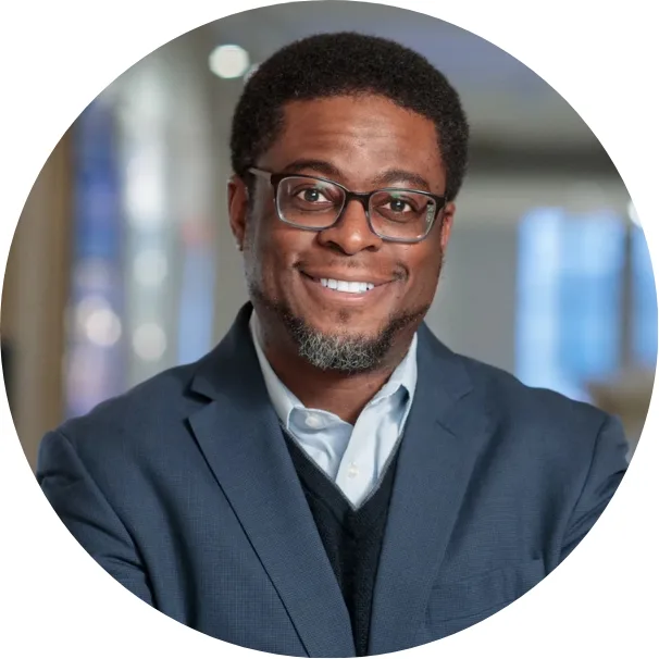 A man with short curly hair, glasses, and a beard smiles while wearing a dark blue suit, light blue shirt, and black sweater. The background is softly blurred with blue and beige tones.