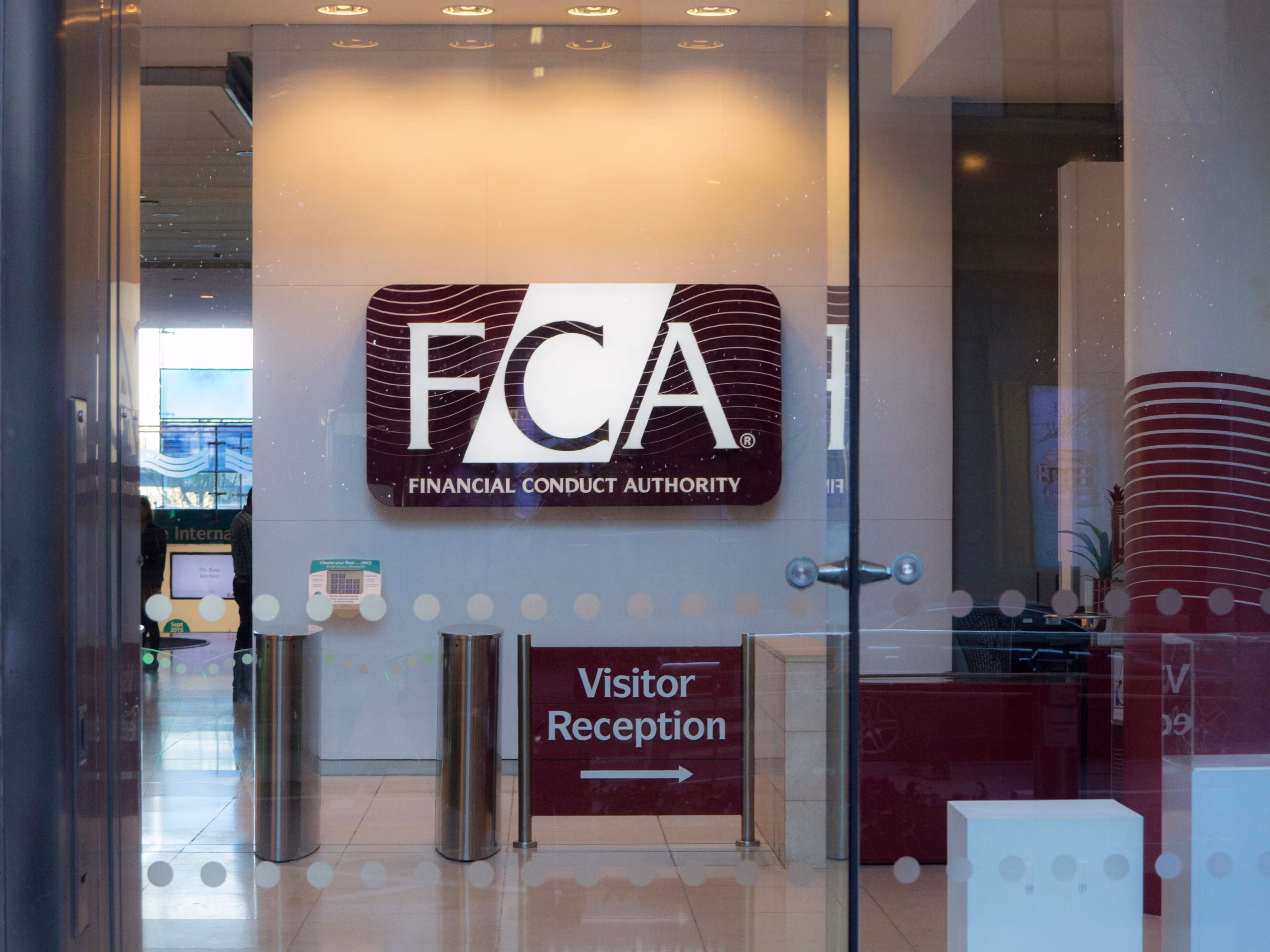 Entrance to the Financial Conduct Authority (FCA) office, showing a large FCA logo on the wall and a sign pointing to Visitor Reception, viewed through glass doors.