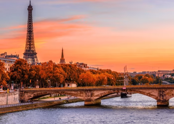 The Eiffel Tower rises above Parisian buildings and autumn trees at sunset, with a stone bridge crossing the Seine River in the foreground under a colorful orange and pink sky.