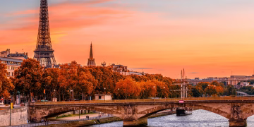 The Eiffel Tower rises above Parisian buildings and autumn trees at sunset, with a stone bridge crossing the Seine River in the foreground under a colorful orange and pink sky.