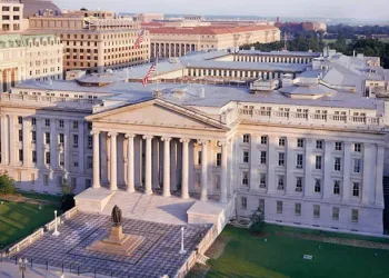 Aerial view of the U.S. Treasury Building in Washington, D.C., featuring neoclassical architecture, tall columns, a statue in front, green lawns, and surrounding government buildings under a clear sky.