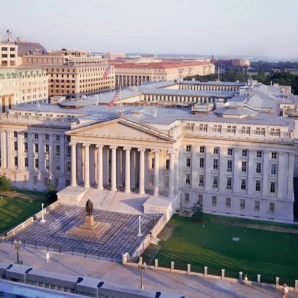 Aerial view of the U.S. Treasury Building in Washington, D.C., featuring neoclassical architecture, tall columns, a statue in front, green lawns, and surrounding government buildings under a clear sky.