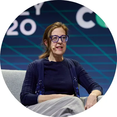 A woman with glasses and a headset microphone sits on a gray chair, speaking on stage with Team CCI. The geometric blue and green background and partial event text are visible behind her.
