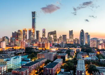 A city skyline at sunset featuring modern skyscrapers and distinctive high-rise buildings, with residential and commercial areas in the foreground and a colorful sky above.