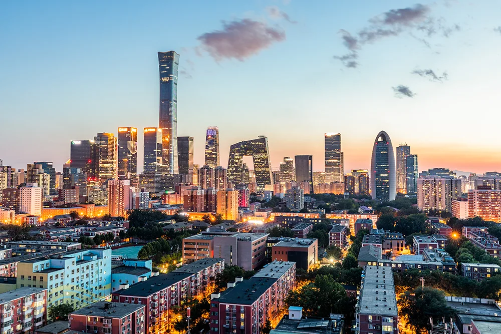 A city skyline at sunset featuring modern skyscrapers and distinctive high-rise buildings, with residential and commercial areas in the foreground and a colorful sky above.
