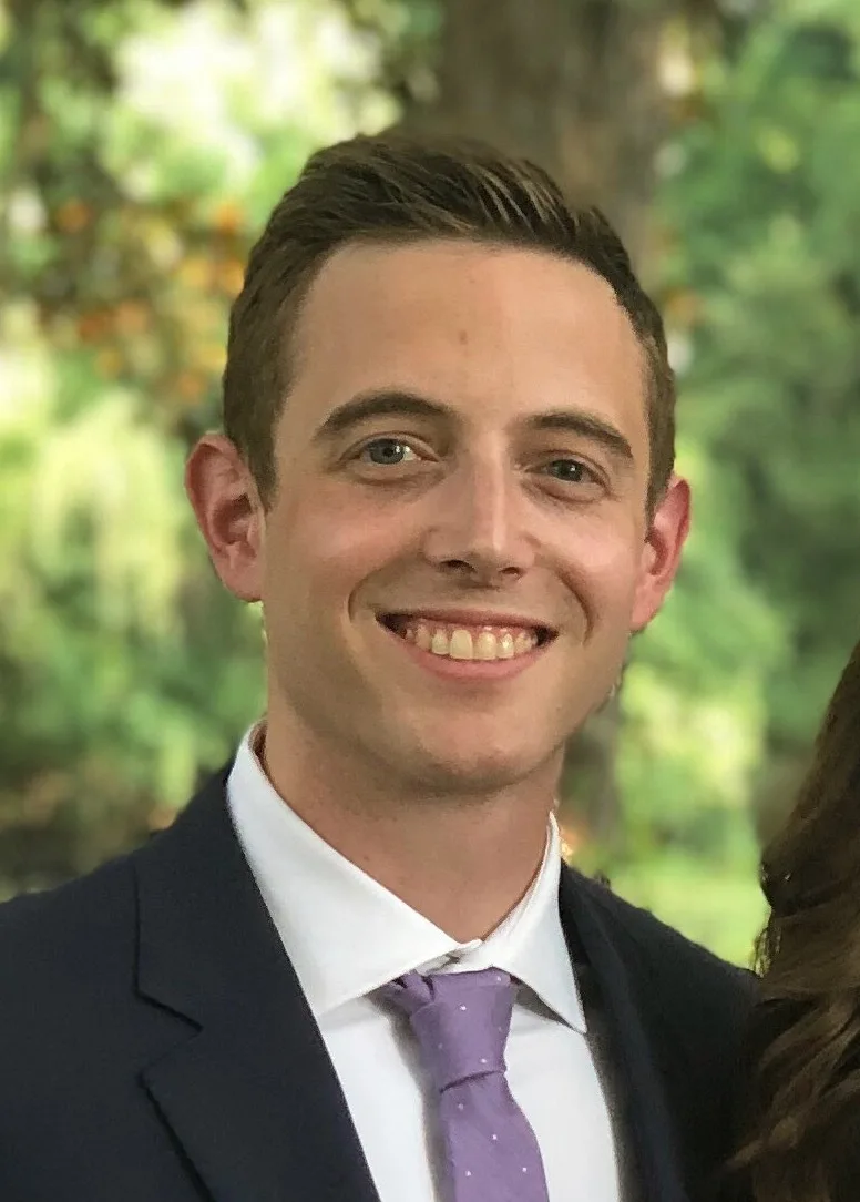 A man in a dark suit, white shirt, and light purple tie smiles at the camera in an outdoor setting with green trees blurred behind him, representing Team CCI.