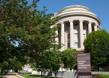 A large, round, neoclassical stone building with columns is surrounded by trees. In front is a sign reading Federal Trade Commission Building with directions to the visitor entrance. The sky is clear and blue.