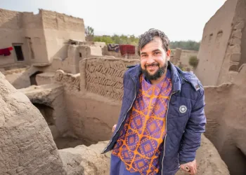 A man wearing a blue jacket and orange patterned shirt stands smiling among mud-brick buildings and walls in a rustic village setting, with rugs hanging in the background.
