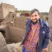 A man wearing a blue jacket and orange patterned shirt stands smiling among mud-brick buildings and walls in a rustic village setting, with rugs hanging in the background.