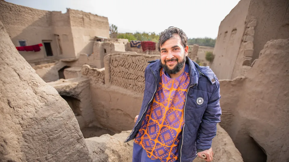A man in a patterned orange and purple outfit and a blue jacket stands smiling among mud-brick buildings in a traditional village setting, with rugs hanging in the background.