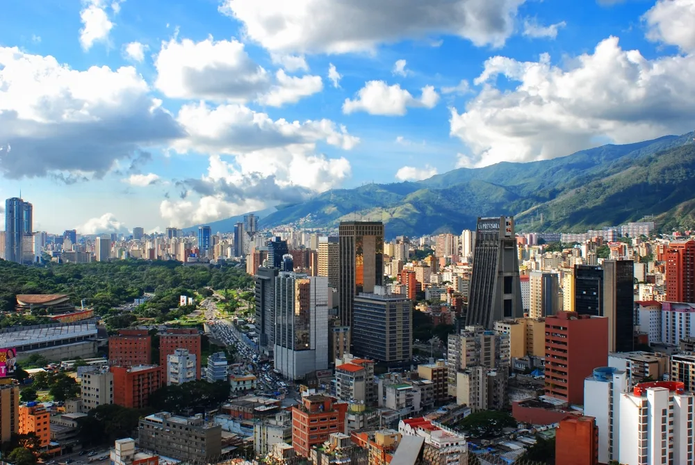 A vibrant city skyline in crypto-driven Venezuela, with tall modern buildings and lush green spaces set against mountains under a blue sky with scattered clouds.