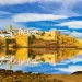 A historic city with white buildings and a prominent golden-brown fortress sits on a rocky shore, reflected clearly in calm water under a blue sky with scattered clouds.