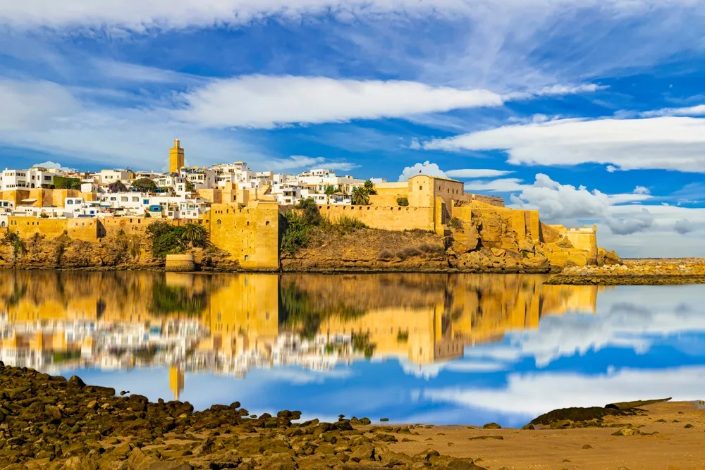 A historic city with white buildings and a prominent golden-brown fortress sits on a rocky shore, reflected clearly in calm water under a blue sky with scattered clouds.