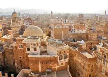 A view of an old city filled with traditional brown brick buildings, domes, and minarets, featuring intricate white detailing, with mountains faintly visible in the hazy background.