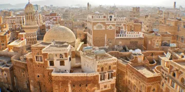 A view of an old city filled with traditional brown brick buildings, domes, and minarets, featuring intricate white detailing, with mountains faintly visible in the hazy background.