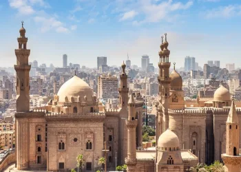 A panoramic view of historic mosques with domes and minarets in Cairo, Egypt, set against a backdrop of modern city buildings under a clear blue sky.