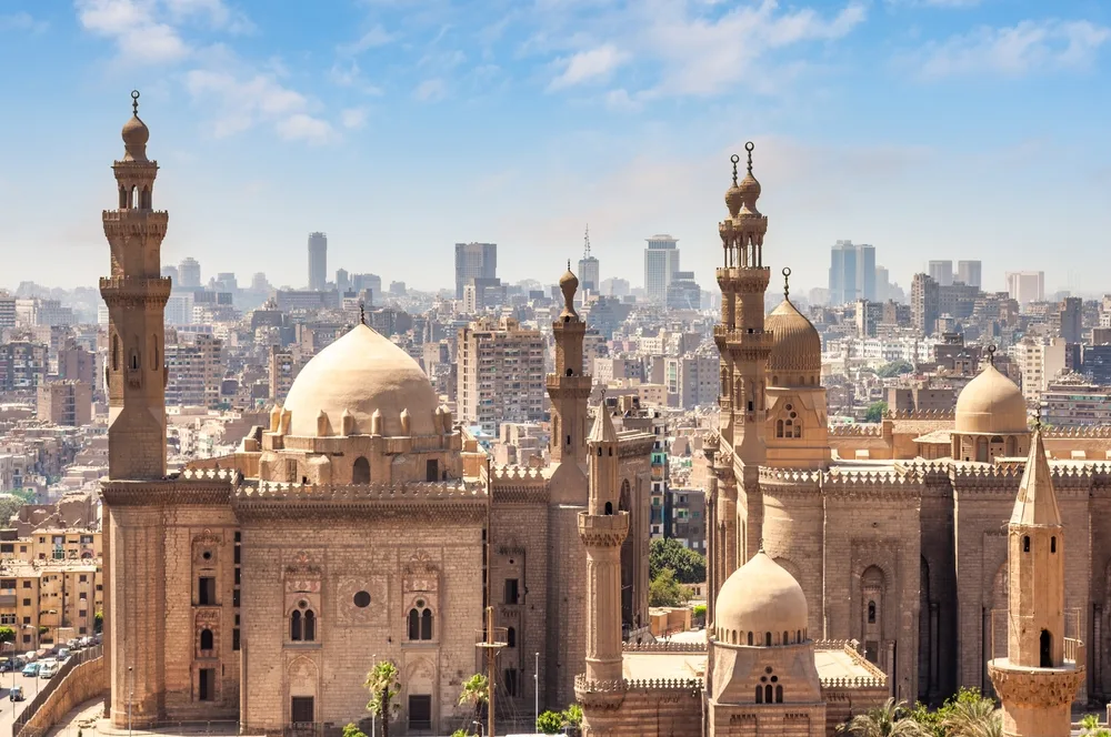 A panoramic view of historic mosques with domes and minarets in Cairo, Egypt, set against a backdrop of modern city buildings under a clear blue sky.