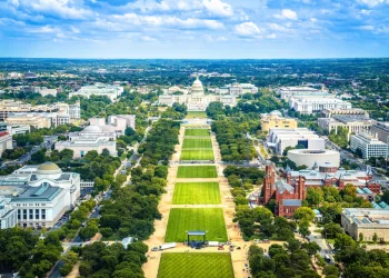 Aerial view of the National Mall in Washington, D.C., showing green lawns, tree-lined paths, and prominent buildings like the U.S. Capitol, museums, and the Smithsonian Castle under a partly cloudy sky.
