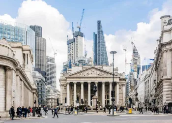 People walk in front of the Bank of England and Royal Exchange in London, surrounded by modern skyscrapers and construction cranes under a partly cloudy sky.
